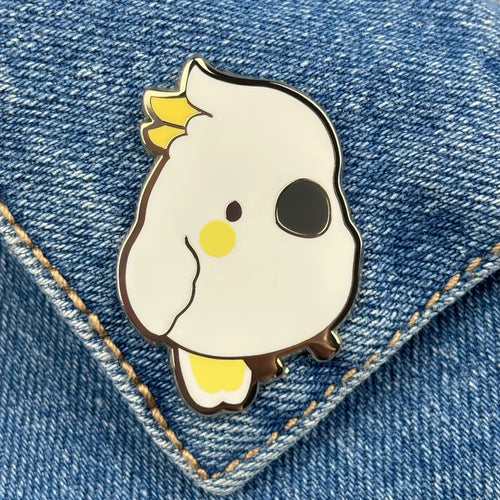 The Sulphur Crested Cockatoo Pin Pinstachiio by Blue Dog Posters is an adorable enamel pin featuring a cartoon cockatoo with yellow cheeks and crest, displayed on blue denim fabric with visible stitching.