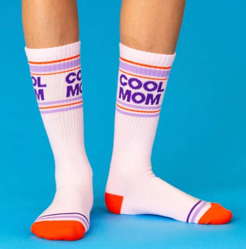 Someone stands on a bright blue background wearing Blue Dog Posters’ Cool Mom Gym Crew Socks—these unisex socks feature white fabric, red toes and heels, purple and orange stripes, and “COOL MOM” in purple on each sock.