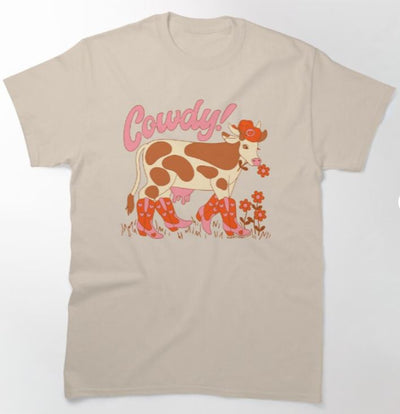 The Cowdy T-shirt Nat MED by Blue Dog Posters features a playful illustration of a cow wearing red boots in a field of flowers. Above the cow, the word "Cowdy!" is written in pink, stylized text. The charming design, drawn in shades of brown, orange, and pink, is available in a natural beige color. Be sure to check the size chart for the perfect fit!