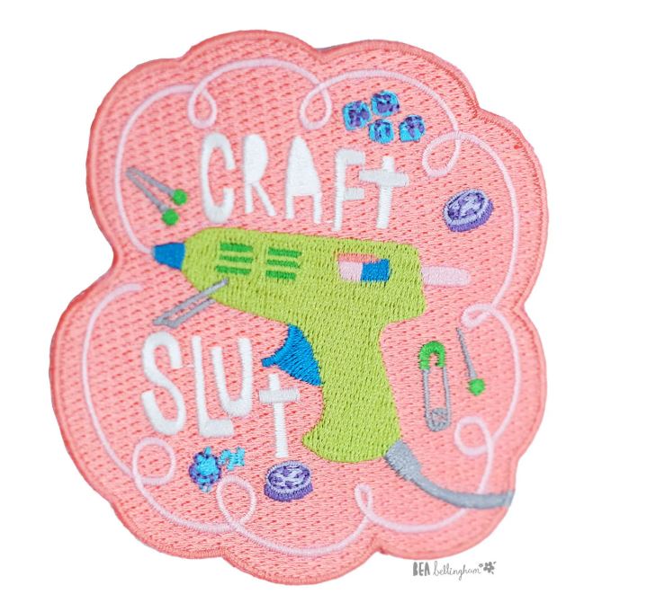 The Craft Slut Patch by Blue Dog Posters is an iron-on embroidered patch with a pink scalloped border, green hot glue gun, craft supplies, and "CRAFT SLUT" in playful white letters—perfect for fans of quirky style.