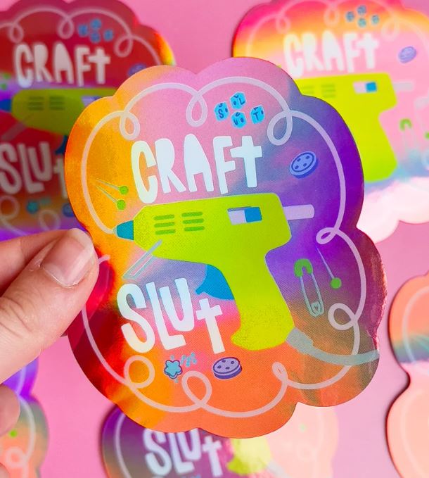 A hand holds the Craft Slut Holographic Sticker by Blue Dog Posters, featuring a green glue gun, bold "CRAFT SLUT" text, sewing icons like buttons and pins, all on a vibrant pink background.