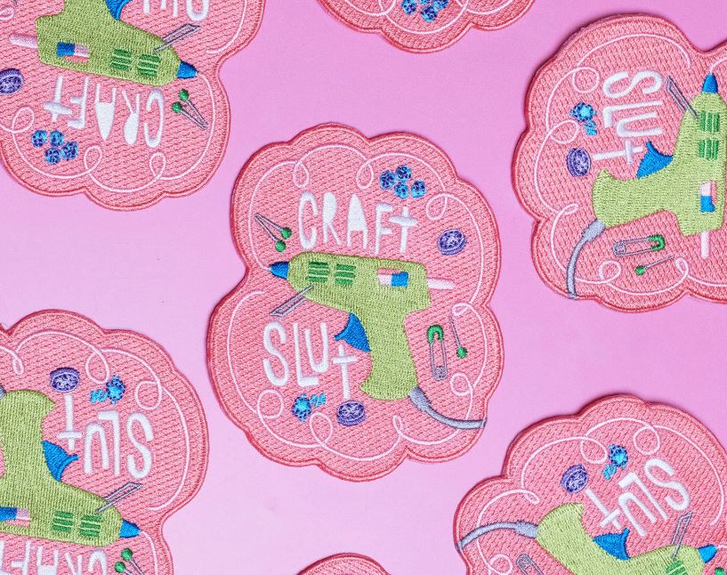 The Craft Slut Patch by Blue Dog Posters features a green glue gun, blue buttons, and "CRAFT SLUT" in white and blue on a pink scalloped background. This embroidered iron-on patch repeats the design on pink.