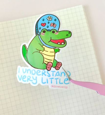 The I Understand Very Little Vinyl Single Sticker by Jess Chen (Blue Dog Posters) features a smiling cartoon alligator in a blue helmet with "I UNDERSTAND VERY LITTLE" below, perfect as a waterproof laptop decal.