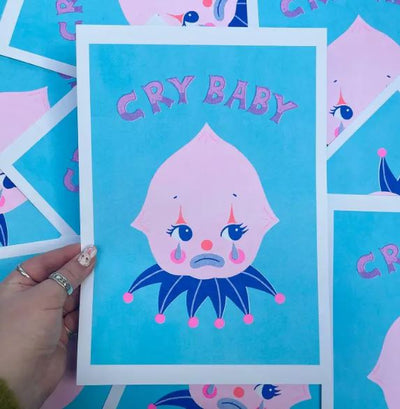 A hand holds the Cry Baby A4 Risograph by Amy Hastings from Blue Dog Posters, featuring a cartoon character with a pink face and teardrops on a blue background. The character has a dark collar with pink dots and a sad expression, with "Cry Baby" printed above in decorative letters.