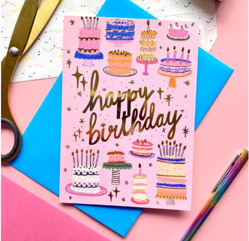 The Happy Birthday Cake Pattern Greeting Card by Blue Dog Posters features colorful cake illustrations and gold foil “happy birthday” lettering on pink, shown with a blue envelope amid scissors, pens, and confetti.