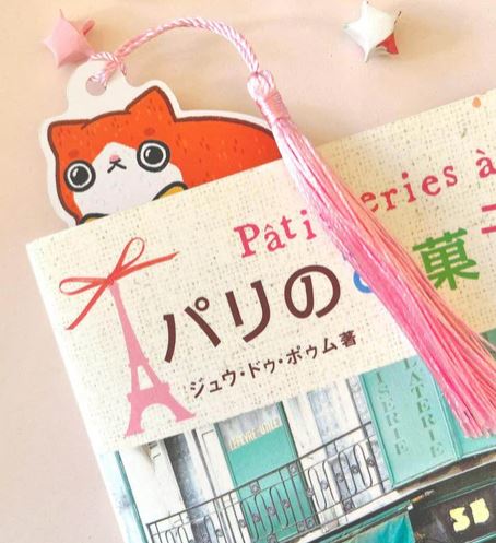 The Bookmark Cat Stack by Jess Chen from Blue Dog Posters, featuring an orange cat with a pink tassel, is shown in a book with Japanese and French text, accompanied by two origami stars—one white and one pink.
