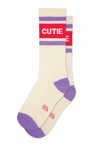 The Cutie Gym Crew Socks by Blue Dog Posters are cream-colored unisex cotton socks featuring purple toes and heels, purple and red stripes, and "CUTIE" in bold white letters on a red band for a playful touch.