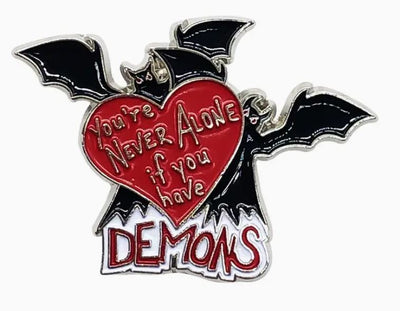The "You're Never Alone Demons Enamel Pin" by Blue Dog Posters is a funny and unique soft enamel pin featuring two black bats with outstretched wings, holding a red heart that reads "You're never alone if you have demons." The word "DEMONS" is boldly written in uppercase letters at the bottom in red and white.