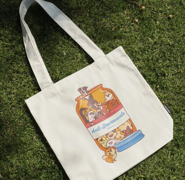 A Blue Dog Posters Anti-Depressants Dog Edition Tote Bag lies on grass, featuring an illustration of a jar filled with happy cartoon dogs and labeled "Anti-Depressants.