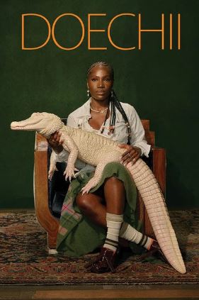 A woman with braided hair sits on a chair against a dark green background, holding a large albino alligator on her lap. Above her, "Doechii Alligator Bites" by MUS 330 is displayed in bold orange letters on this striking large poster.