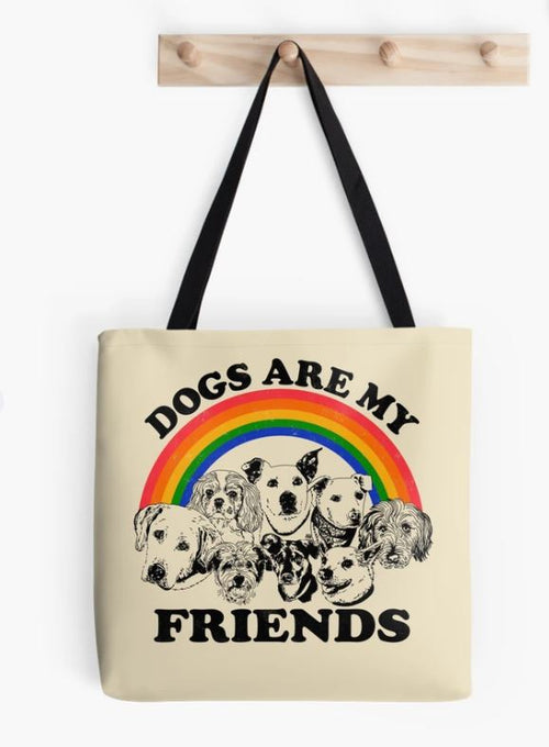 A sublimation-printed tote bag named "Dogs are my Friends" by Blue Dog Posters is hanging on a light wooden rack. It features illustrations of various dog breeds with a rainbow arch above them and the words "Dogs Are My Friends" in black. The bag has black straps and highlights Audrey Herbertson’s distinctive style.