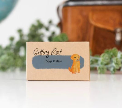 A small rectangular box from Blue Dog Posters, labeled "Getting Lost Cards - Dogs Edition," sits on a white surface. The box, perfect for a spontaneous fun adventure, features an illustration of a yellow dog. In the background, there is a blurred brown bag and green foliage.