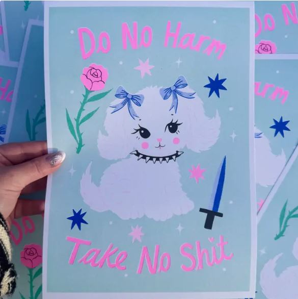 A hand holds the Do No Harm A4 Risograph by Amy Hastings from Blue Dog Posters, showing a white dog with blue bows amid stars, roses, and a blue knife. "Do No Harm, Take No Shit" in pink text. Printed on recycled coffee cup paper.