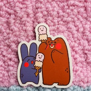 The Double Scoop Vinyl Single Sticker Misu Juju by Blue Dog Posters features a cute purple bunny and brown bear with smiling ice cream cones on a pink crocheted background—adorable and waterproof!.