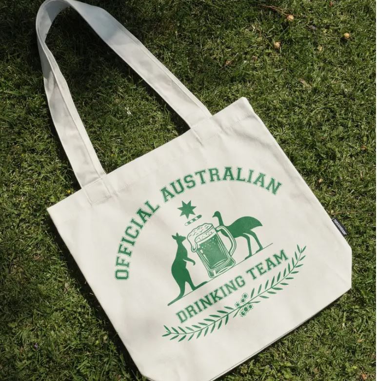 The Official Australian Drinking Team Tote Bag Natural by Blue Dog Posters sits on the grass, crafted from cotton canvas and featuring green text and graphics of a kangaroo, emu, beer mug, and laurel design.