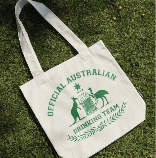 The Official Australian Drinking Team Tote Bag Natural by Blue Dog Posters sits on the grass, crafted from cotton canvas and featuring green text and graphics of a kangaroo, emu, beer mug, and laurel design.