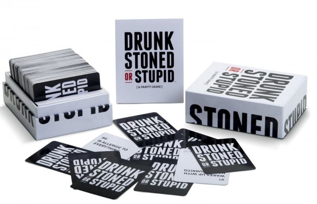 A party game from Blue Dog Posters called "Drunk Stoned or Stupid" is displayed. The image shows the game box, an instruction booklet, and several prompt cards scattered around, each featuring humorous or challenging statements. The box has bold black and red text on a white background.