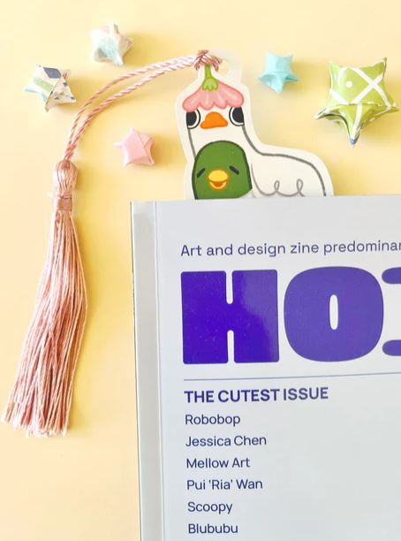 A magazine titled "THE CUTEST ISSUE" with a pastel pink tassel and the Bookmark Duck Stack by Jess Chen from Blue Dog Posters peeking out. Colorful origami stars on a yellow background—ideal for cute stationery and marine life fans.