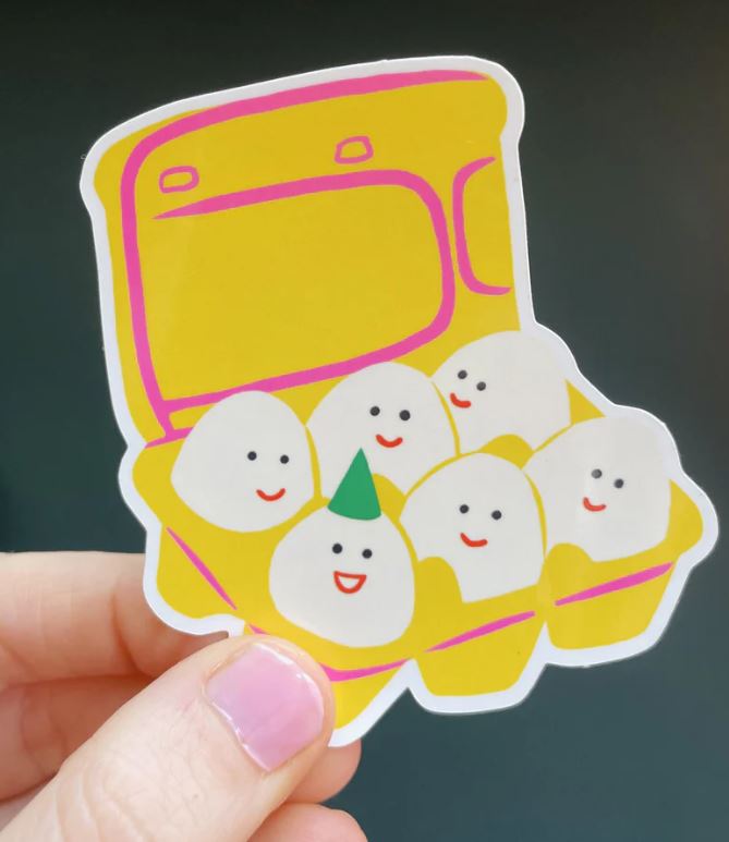 A hand holds the Egg Carton Vinyl Sticker by Blue Dog Posters, featuring five smiling eggs in a yellow carton—one sporting a green party hat—all with cheerful faces.