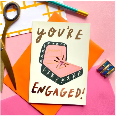 Blue Dog Posters' You're Engaged Ring Greeting Card features bold “You’re Engaged!” text and a ring-in-a-box illustration. This FSC certified card is shown on pink with scissors, pen, colored envelope, and patterned tape nearby.