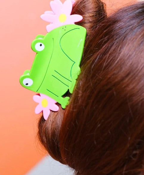 A close-up of brown hair in a bun, secured with Blue Dog Posters’ Princess Frog Hair Claw—a bright green frog-shaped clip adorned with pink flowers, ideal for anyone who loves quirky accessories.