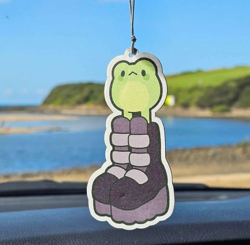 The Goth Froggy Bubblegum Air Freshener by Blue Dog Posters, featuring a green frog in purple boots, hangs from a car’s mirror as a river and blue sky can be seen through the windshield.