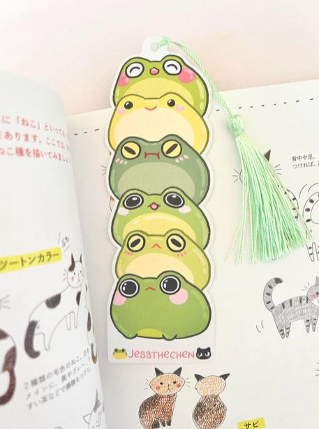 The Bookmark Frog Stack by Jess Chen from Blue Dog Posters is a die-cut bookmark featuring six stacked cartoon frogs in green and yellow, each with unique expressions, made from premium cardstock with a light green tassel.