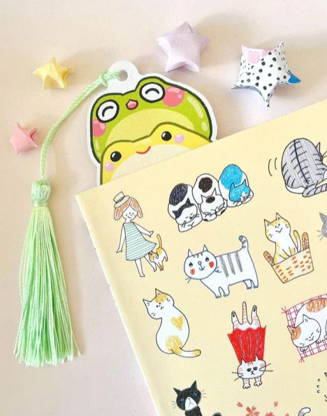 A notebook with adorable cat art, Blue Dog Posters' "Bookmark Frog Stack" by Jess Chen—a green frog die-cut bookmark with a tassel—and colorful origami stars displayed on premium cardstock.