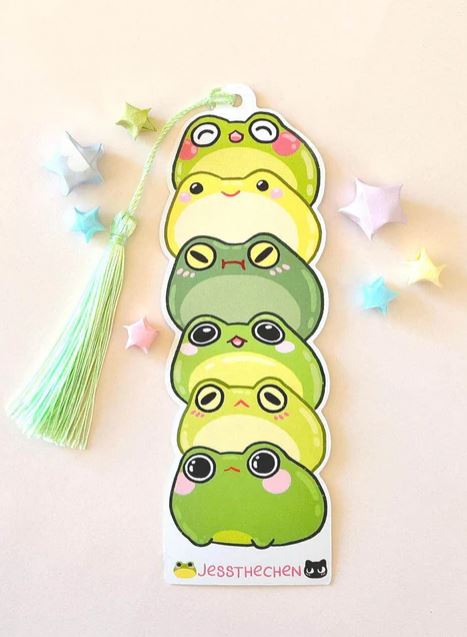 The Bookmark Frog Stack by Jess Chen from Blue Dog Posters is a die-cut bookmark with five green cartoon frogs, each with a unique expression, made of premium cardstock. It has a green tassel and pastel star origami accents.