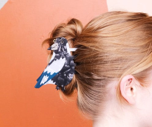 A person with light brown hair in a messy bun wears the Blue Dog Posters Magpie Hair Claw, shaped like a black-and-white bird. The background shows a peach and orange semicircle design.