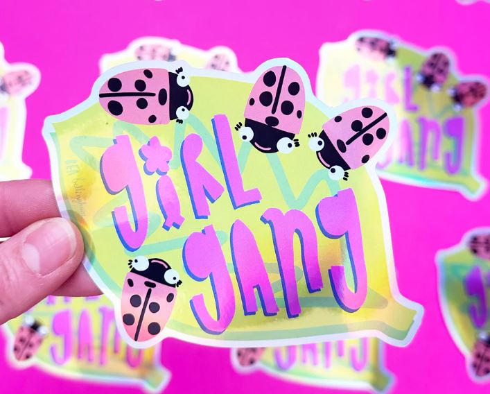 A hand holds the water-resistant Girl Gang Holographic Sticker by Blue Dog Posters, shaped like a green leaf with “Girl Gang” in bold colors and pink ladybugs. Similar stickers are scattered on a bright pink surface.