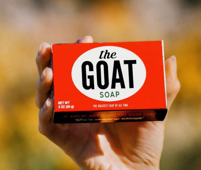 A hand holds a red and white box labeled "GOAT Triple Milled Boxed Bar Soap" by Blue Dog Posters, featuring retro-inspired packaging, against a blurred yellow background.