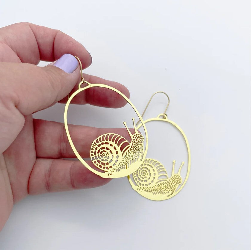 A hand holds a "Snails in Gold Dangle Earrings" by Blue Dog Posters, highlighting the whimsical jewelry with its intricate gold metal design. Another statement earring lies on a white surface, while light purple nails add a playful touch to the scene of charmingly slippery snails.