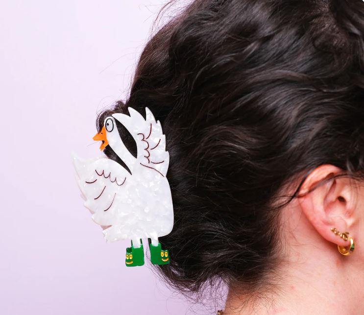 A person with dark, curly hair wears a large, white Silly Goose Hair Claw by Blue Dog Posters, decorated with green shoes. Their ear is visible with two small gold hoops. The background is light pink.