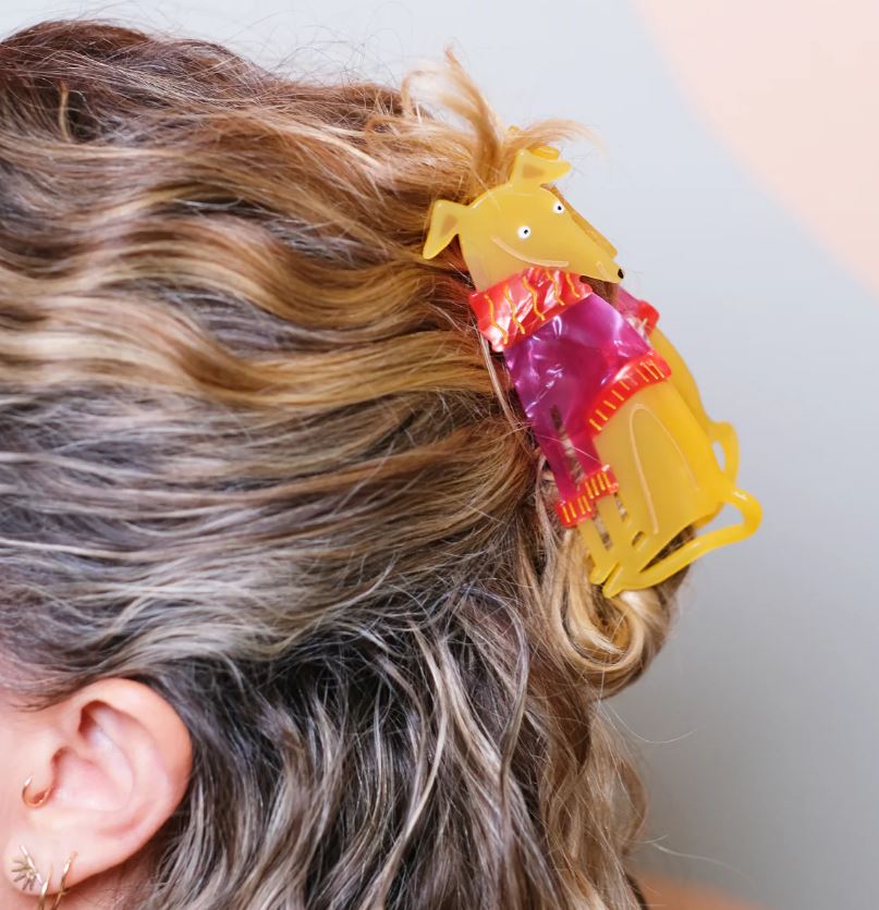 A person with wavy, highlighted hair wears Blue Dog Posters’ Grey Hound in a Jumper Hair Claw, a plant-based acetate accessory shaped like a yellow dog in a cute jumper, with the dog's head and body forming the main structure.