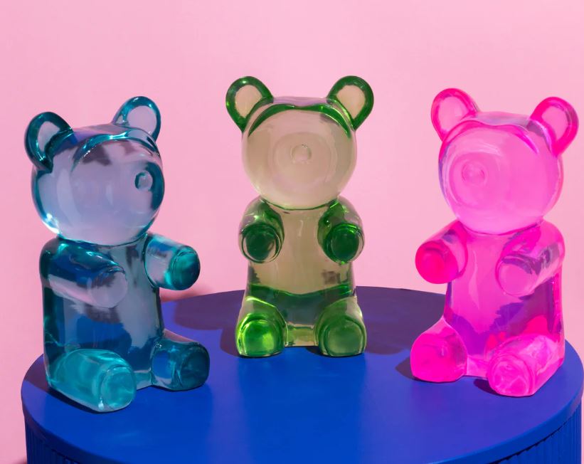 The Barry Bear Blue Resin sculpture by Blue Dog Posters sits on a round blue table against a pink background, adding playful gummy bear decor that also doubles as a unique paperweight.