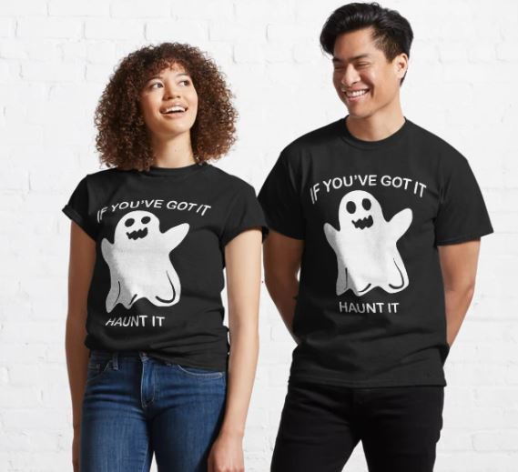 Two people wearing matching Blue Dog Posters Ghost Haunt It T-shirts in black, each featuring a cartoon ghost and the text "IF YOU'VE GOT IT, HAUNT IT." They are standing against a white brick wall, smiling and looking at each other. The person on the left has curly hair and is wearing blue jeans, while the person on the right has straight hair and is also wearing blue jeans.