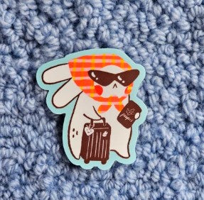 The Holiday Mode Vinyl Single Sticker Misu Juju by Blue Dog Posters features a cartoon bunny with sunglasses and an orange checkered scarf, pulling a suitcase—ideal as a waterproof sticker for travel gear.