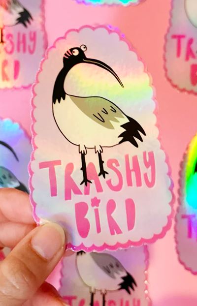 A hand holds the Blue Dog Posters Ibis Trash Bird Holographic Sticker, featuring a cartoon bin chicken and "TRASHY BIRD" in pink. Identical stickers are blurred in the background.