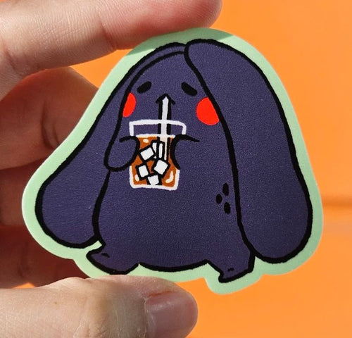 A hand holds the Bunny Iced Coffee Vinyl Single Sticker Misu Juju by Blue Dog Posters—a die-cut waterproof sticker featuring a cute, chubby purple bunny sipping iced coffee, against a vibrant orange background.