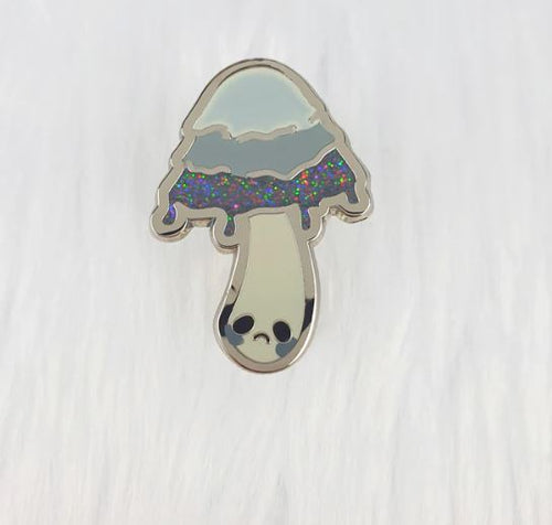 This enchanting enamel pin from Blue Dog Posters, the Inky Cap Mushroom Pin Pinstachio, showcases a whimsical design of an inky cap mushroom with a glittery, multicolored top and a sad face on its stem, all elegantly displayed against a textured white background.