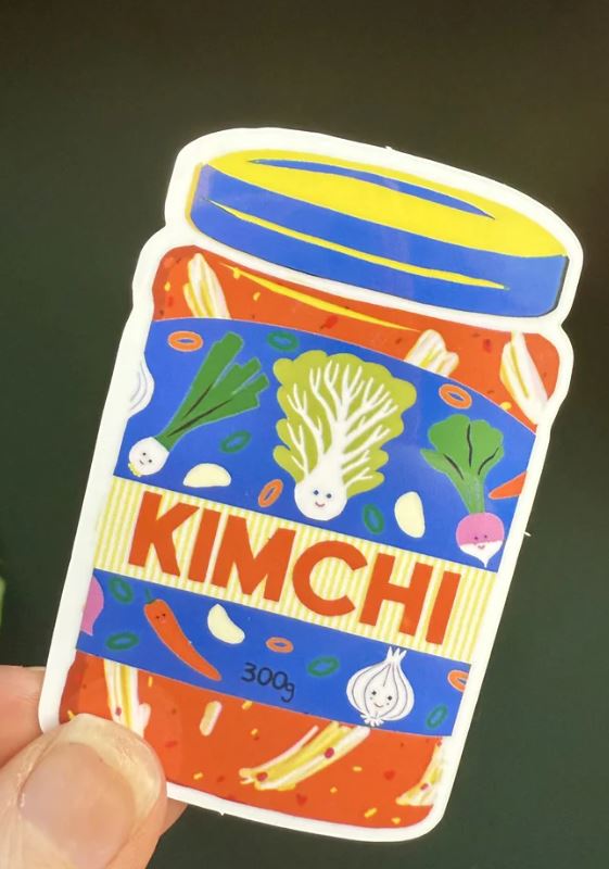 A hand holds the Kimchi Sticker by Blue Dog Posters, featuring cartoon vegetables, the text "KIMCHI," and a "300g" label on a colorful jar with a yellow and blue lid, illustrated by Bristol artist Naomi Wilkinson.