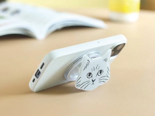 A smartphone with a white case is propped up by the Blue Dog Posters Magnetic Phone Grip Cats - Lucky Cat White, featuring a simple black cat face design. An open book and a yellow item are blurred in the background.