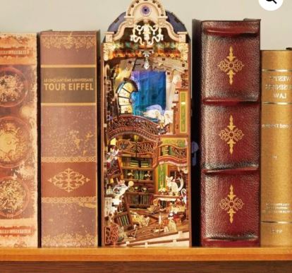 A bookshelf displays several decorative books, with the Magical Library Large Book Nook by Blue Dog Posters nestled between them—a whimsical DIY diorama unveiling an enchanting miniature library scene.