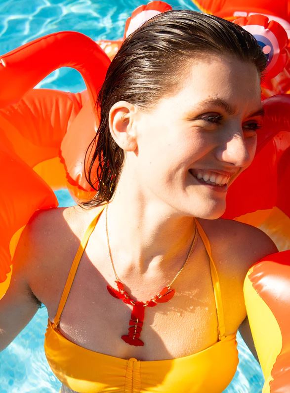 A smiling person in a yellow swimsuit stands in a pool, surrounded by a red inflatable toy. Their wet hair glistens in the sunlight, perfectly complementing the Blue Dog Posters Lobster Necklace - Ruby Shimmer they wear, creating a cheerful atmosphere filled with vibrant ruby hues.
