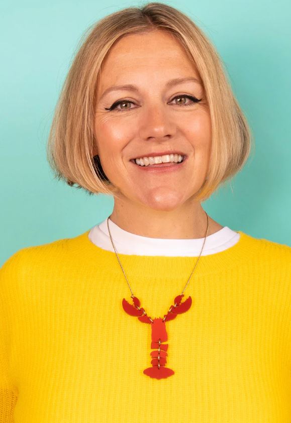 In front of a teal backdrop, a woman radiates joy while wearing a vibrant yellow sweater paired with the Lobster Necklace - Ruby Shimmer by Blue Dog Posters.