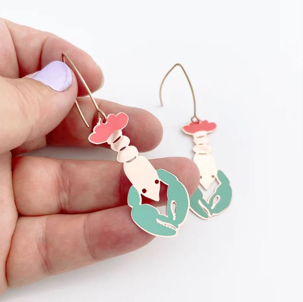 A hand holds a Lobster in Coral Denz Earring by Blue Dog Posters—colorful, lightweight enamel jewelry ideal for seafood lovers. The earring features coral hues; a matching piece is blurred in the background.