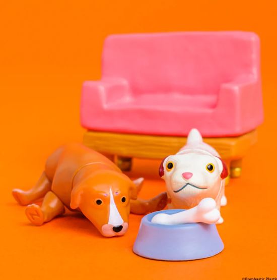 Two collectible mini figures from the "Hello in the World Vol 7 Neil Ewing Blind Box" by Blue Dog Posters rest on an orange surface near a pink toy couch, with one lying down and the other standing by a blue bowl holding a white bone.