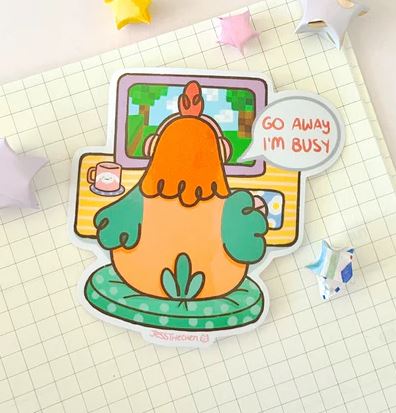 The Go Away Chicken Vinyl Single Sticker by Jess Chen (Blue Dog Posters) features Macy the Chicken at a computer with a coffee, saying "GO AWAY I'M BUSY." A waterproof vinyl sticker for quirky laptops—set on grid notebook and origami stars.