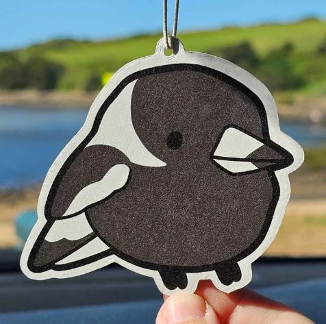 A hand holds the Blue Dog Posters Magpie Grape Air Freshener, featuring a black and white cartoon magpie design, with a blurred outdoor scene of grass, hills, and water in the background.